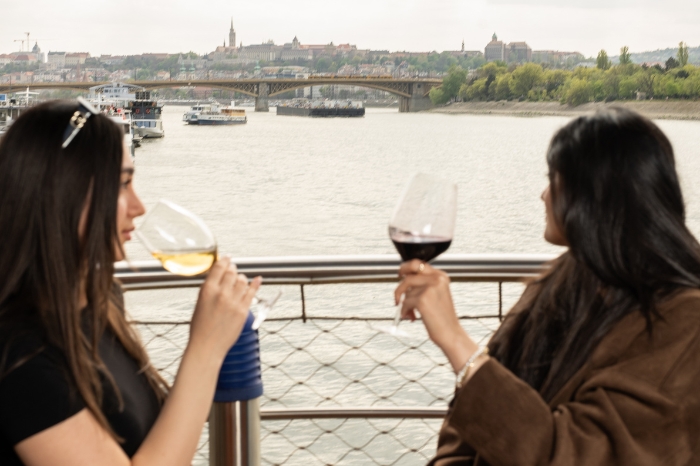 Two women toasting with red and white wine on the deck of the Vogue Boat Restaurant, with the stunning Budapest panorama and Buda Castle in the background.